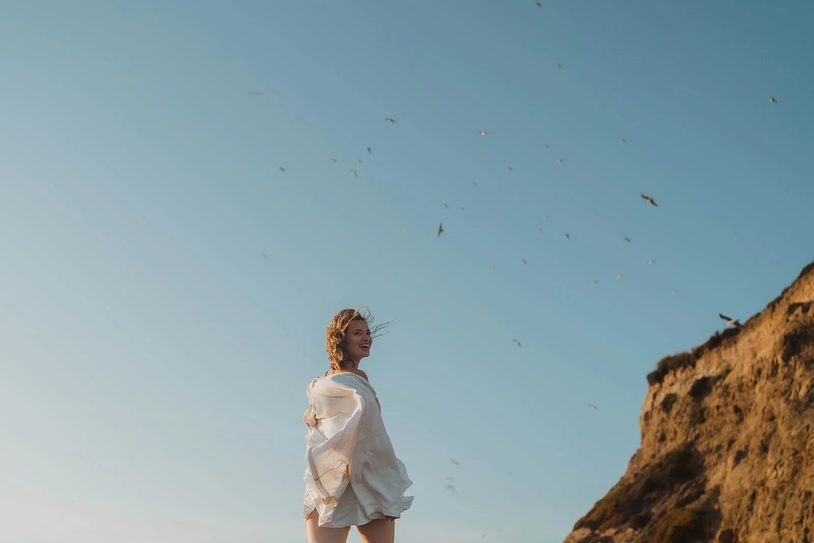 A woman with curly hair standing outdoors near a rock formation, smiling, with a flock of birds in the sky.