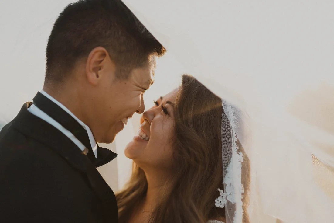 A bride and groom smiling joyfully under a veil, close-up portrait.
