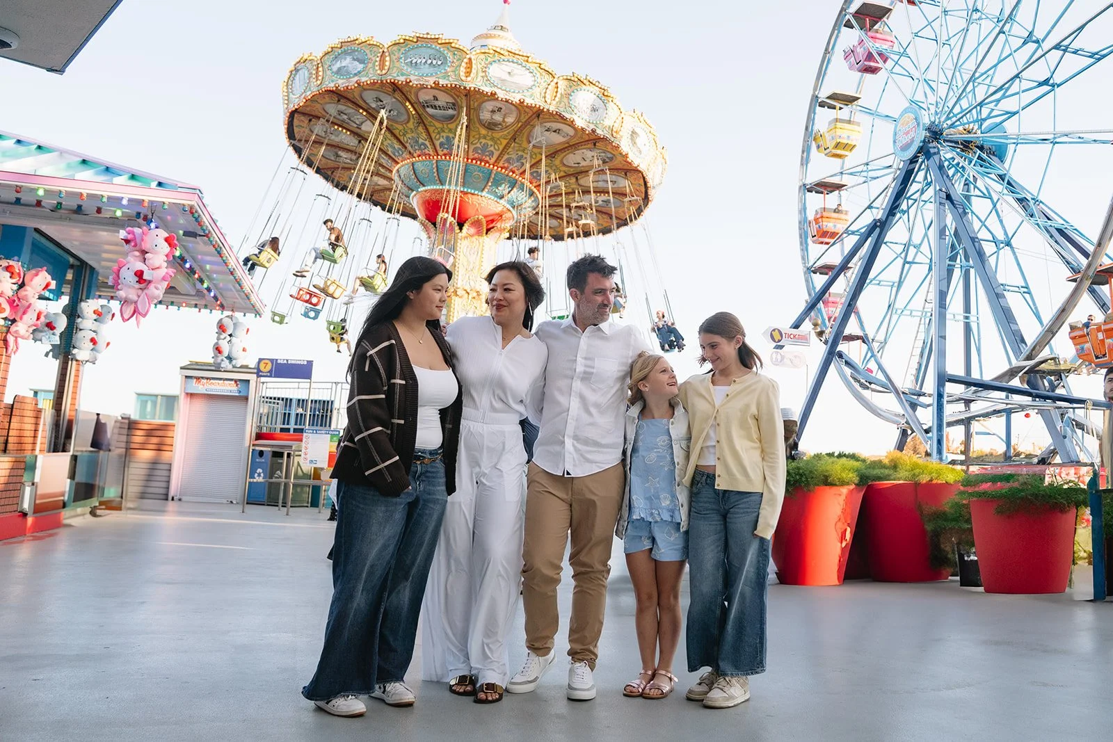 Group of six people smiling and posing together at a carnival, with a large Ferris wheel and a vintage carousel in the background.