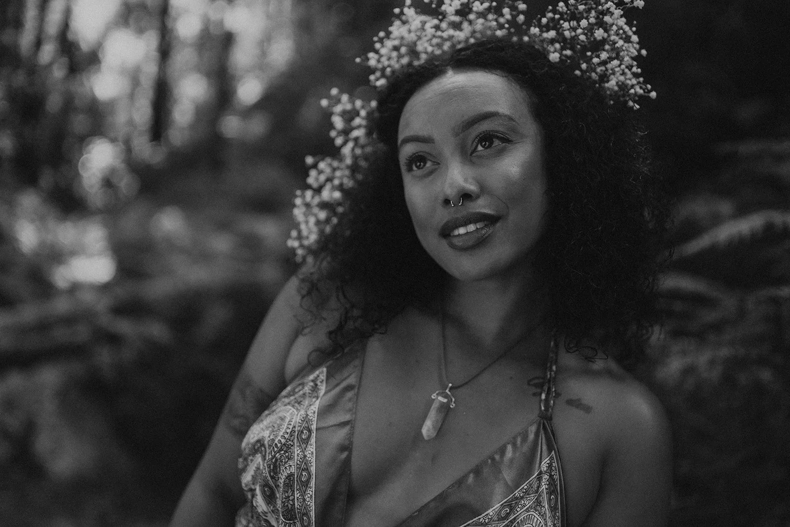 A woman with curly hair and a flower crown is outdoors, smiling and looking slightly away from the camera, wearing a patterned top and a necklace with a crystal pendant.