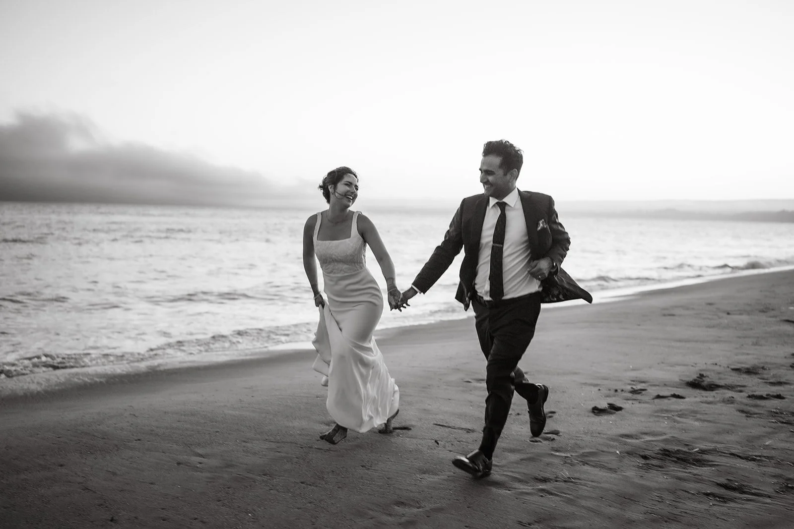 A couple dressed in wedding attire running hand-in-hand on the beach at sunset, smiling and looking at each other.