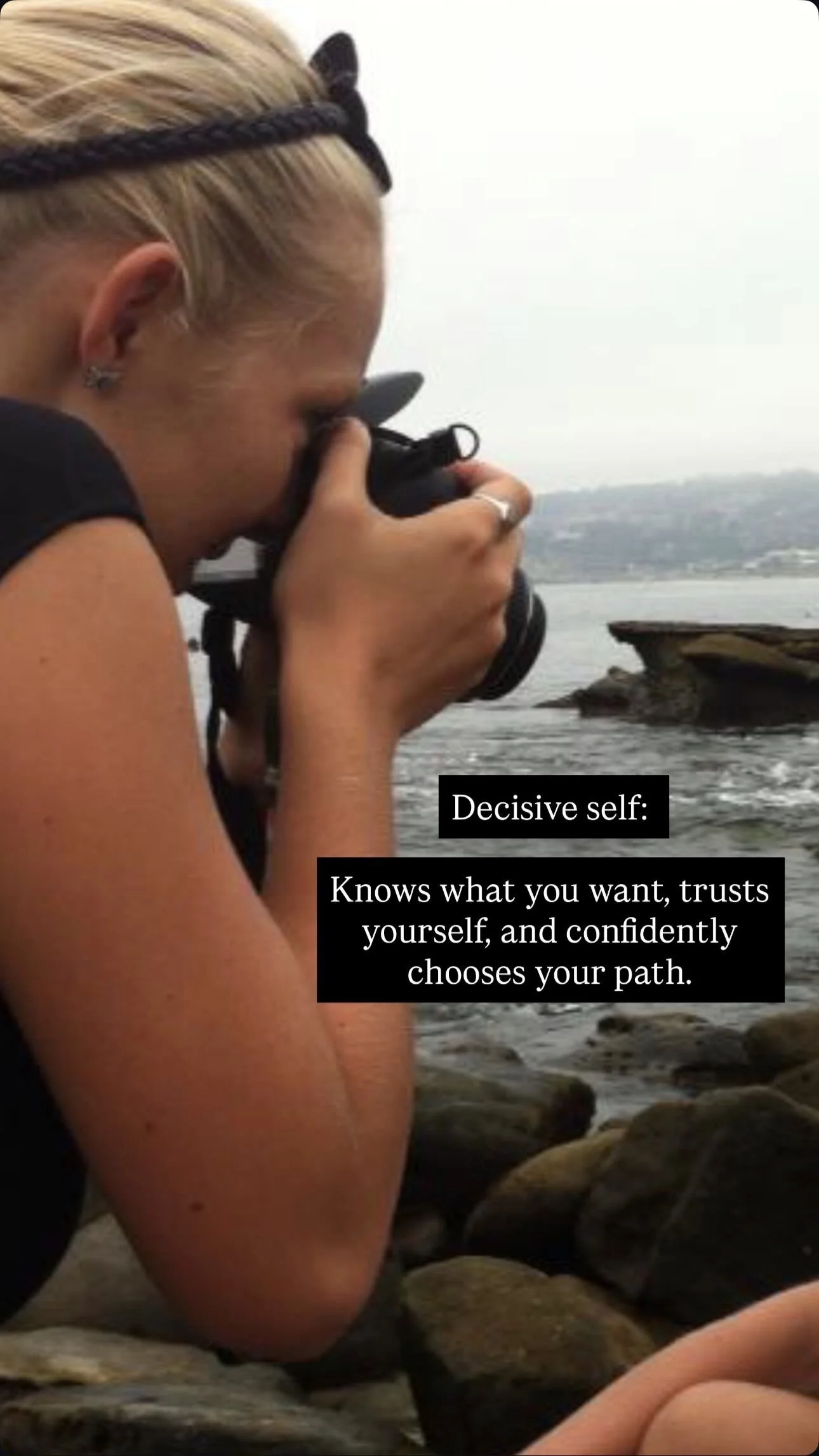 A young woman with blonde hair and earrings taking a photograph near a rocky shoreline with a body of water in the background. Text on the image reads: 'Decisive self: Knows what you want, trusts yourself, and confidently chooses your path.'
