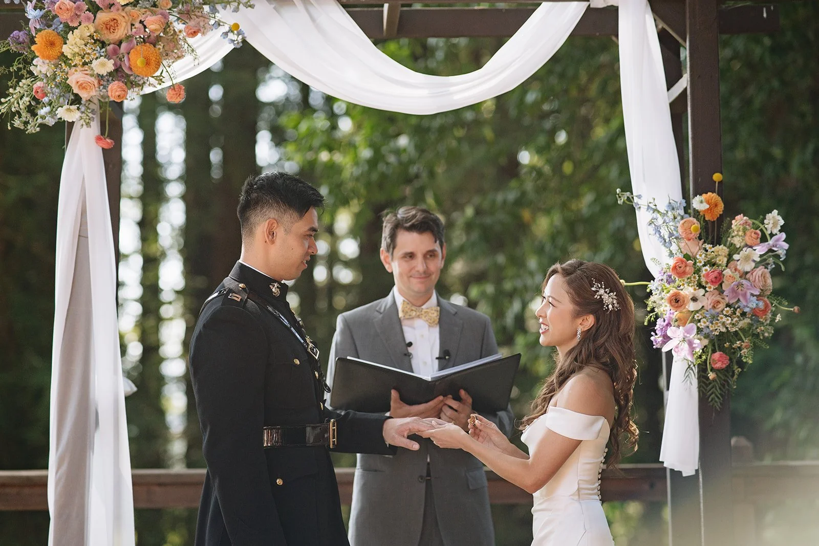 A couple getting married in an outdoor ceremony, exchanging vows under a floral arch, with an officiant standing behind them.
