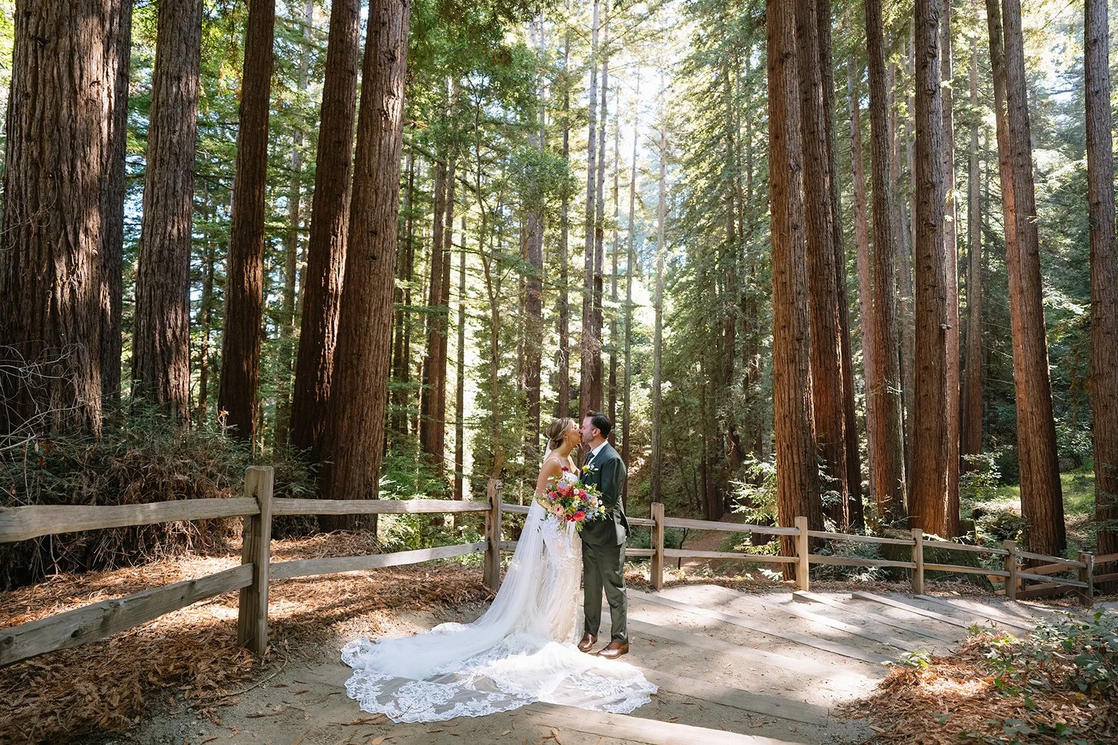 A bride and groom standing close together in a wooded forest, sharing a kiss during their wedding. The bride is dressed in a lace wedding gown with a long train, holding a bouquet of colorful flowers. The groom is in a suit, wearing glasses. Tall trees surround them, filtering sunlight through the leaves, creating a natural and serene atmosphere.