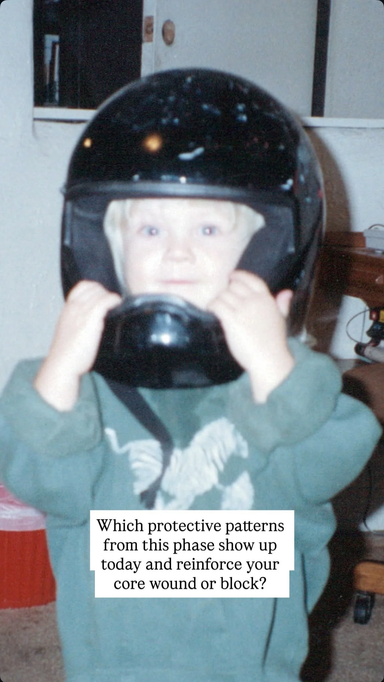 A young child with blonde hair holding a black motorcycle helmet in front of their face, with their face visible through the helmet's visor, inside a room with a white wall and some furniture.