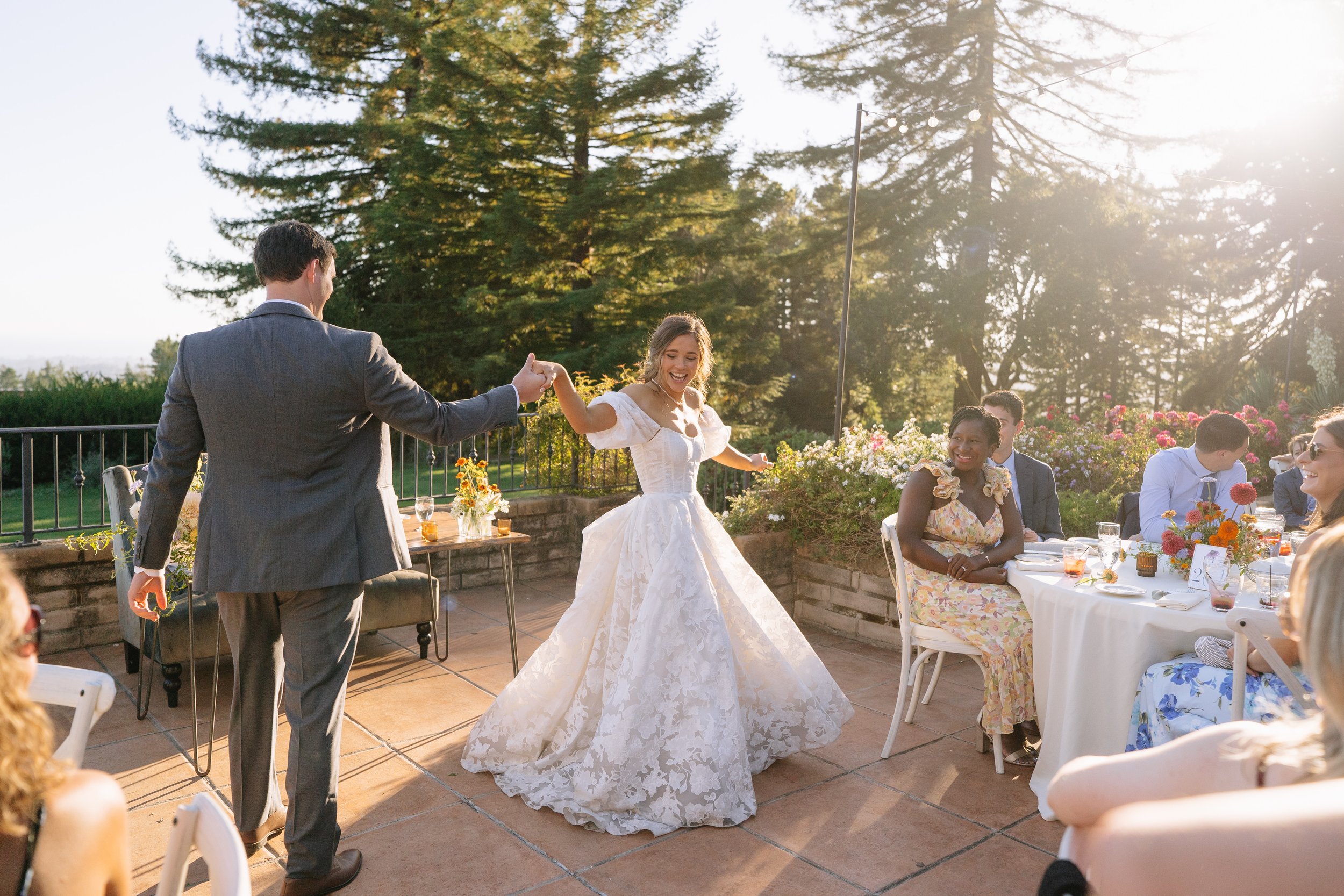 A bride and groom dancing at a wedding reception outdoors in the evening sunlight, with guests sitting at tables with colorful flowers.