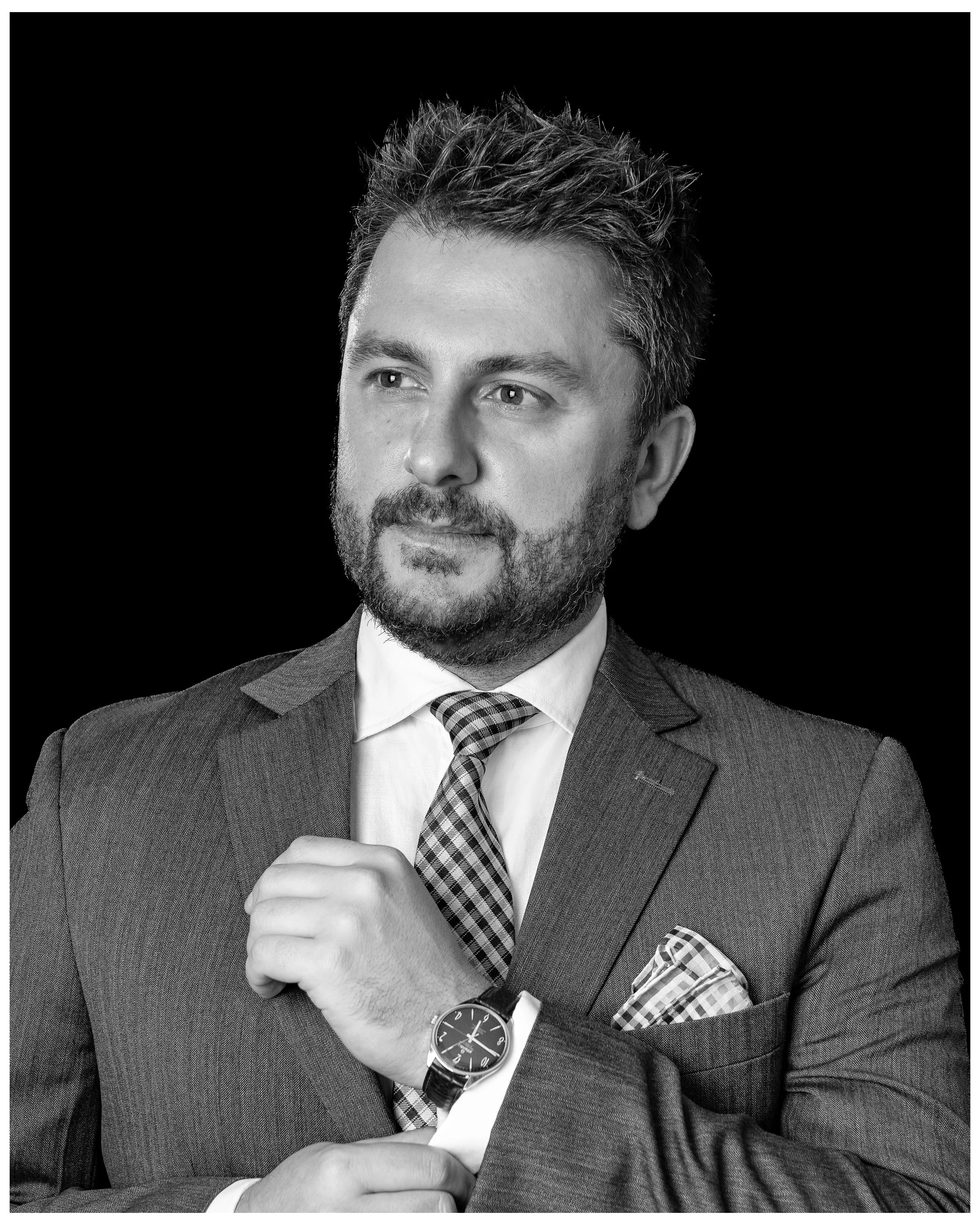 Black and white portrait of a man in a suit, looking to the side, with a watch on his left wrist, adjusting his jacket, against a black background.