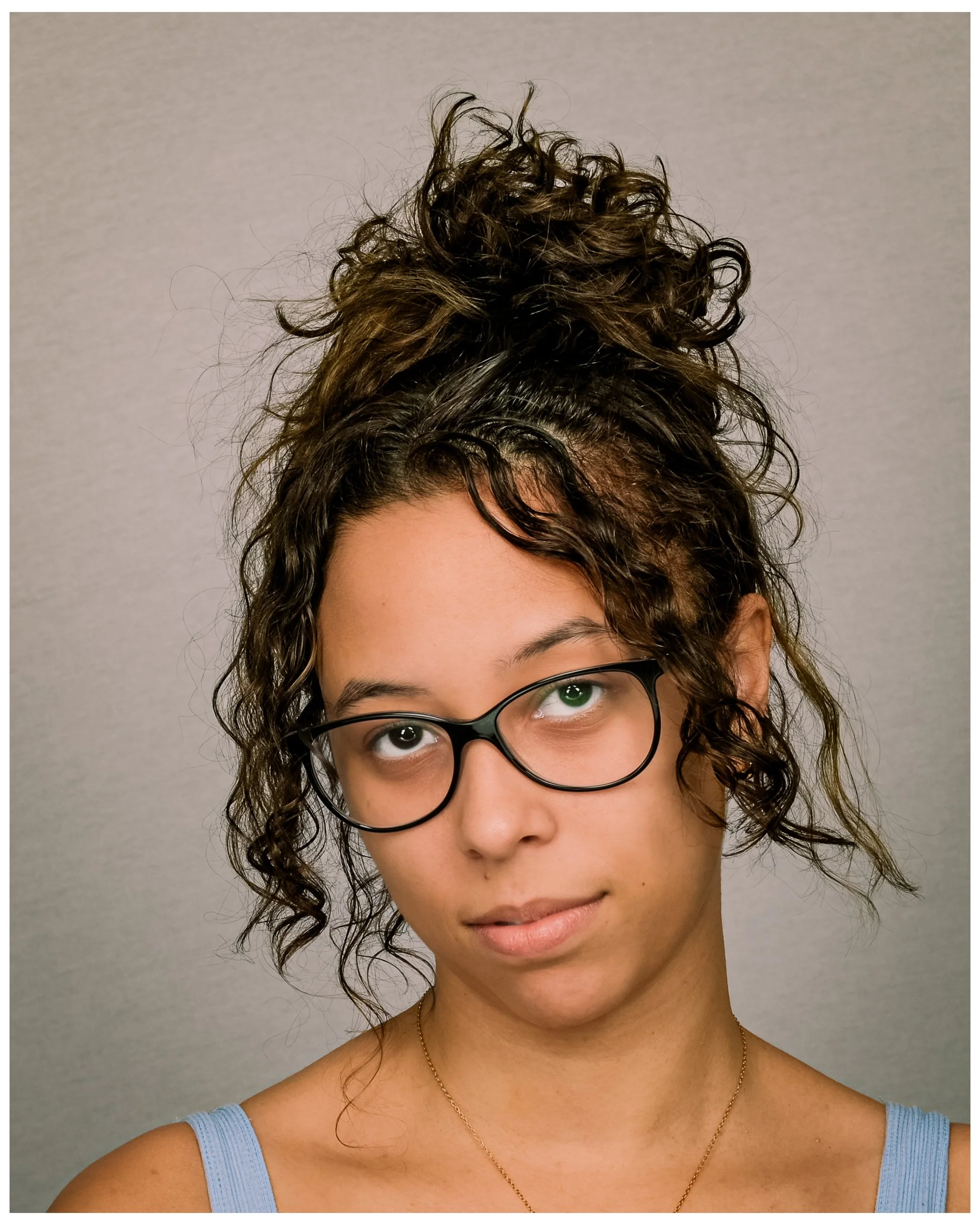 Portrait of a young woman with curly hair styled in a high bun, wearing glasses with black frames, a light blue tank top, and a gold necklace, standing against a plain background.