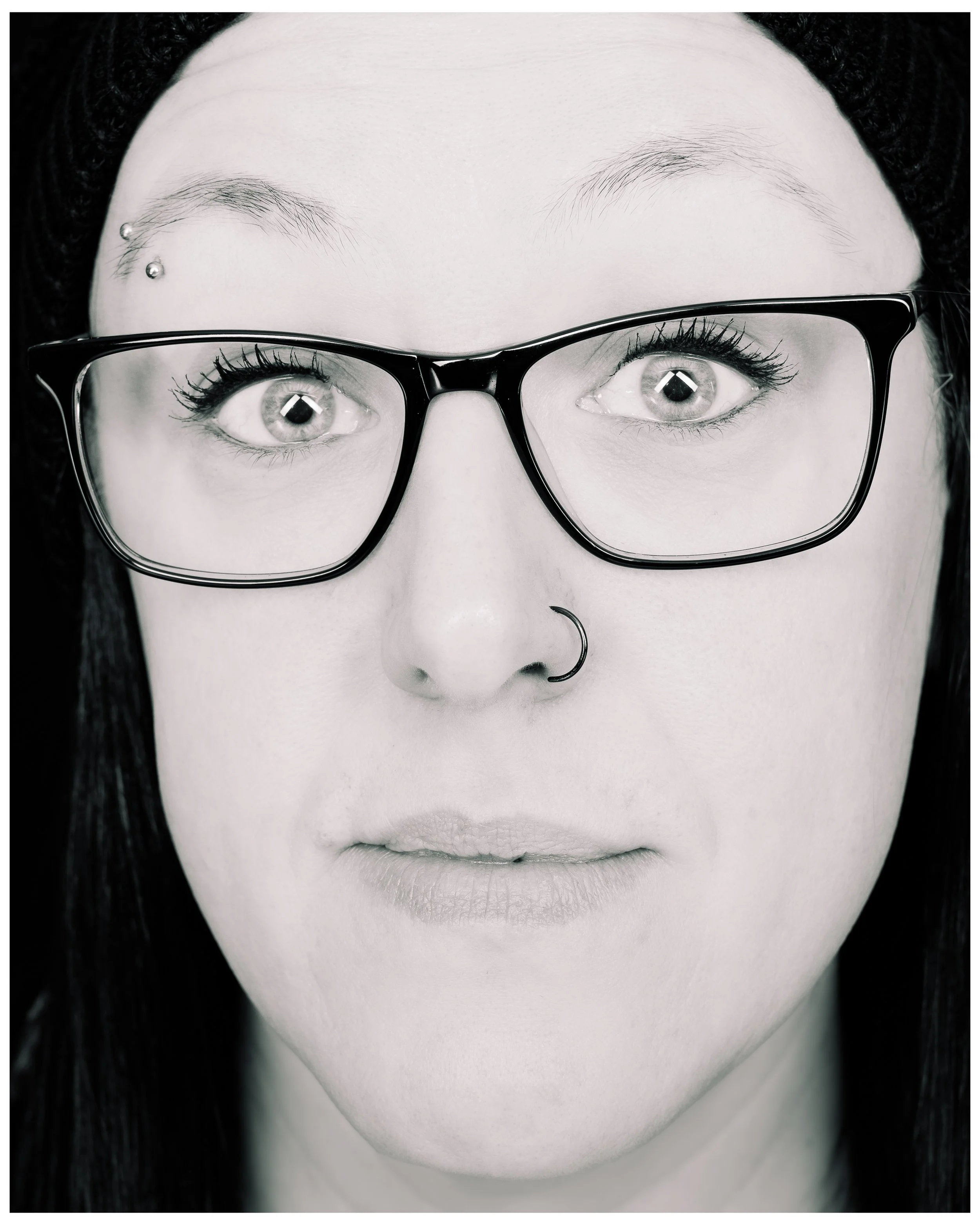 A close-up black and white portrait of a woman wearing glasses, with two facial piercings: one on her eyebrow and one on her nose, and with long dark hair.