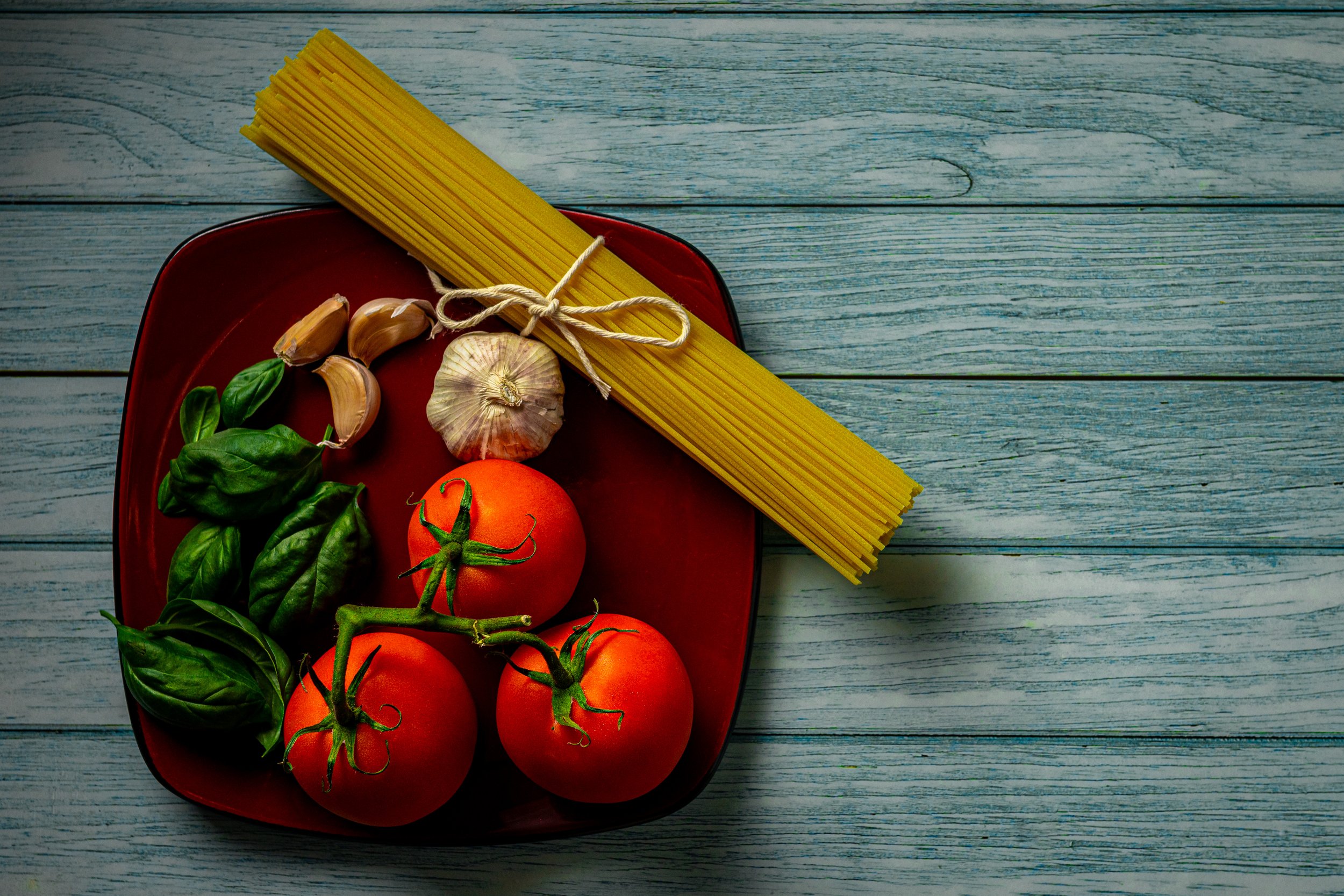 Fresh tomatoes, basil, garlic cloves, and spaghetti on a red plate with a rustic wooden background.