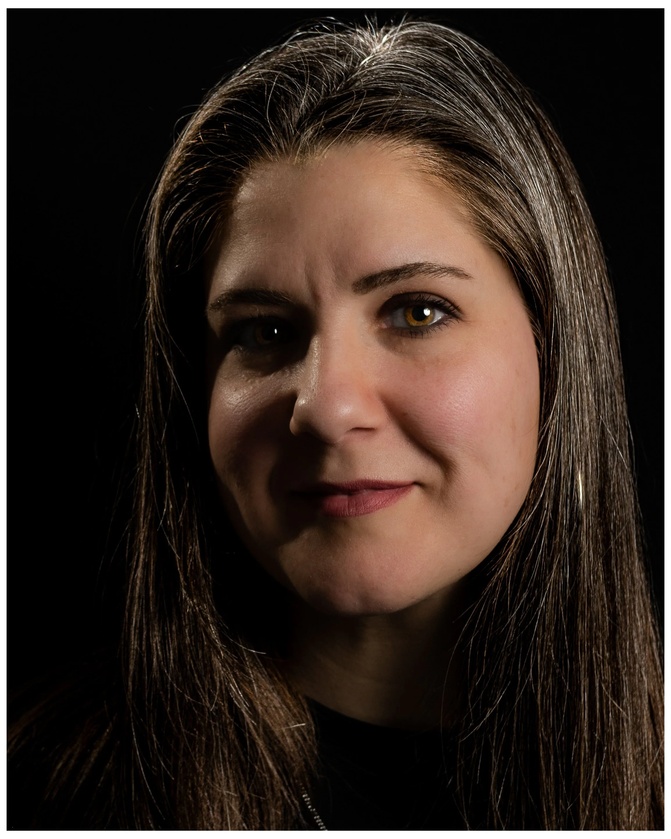 Close-up portrait of a woman with long brown hair and hazel eyes, smiling softly against a black background.