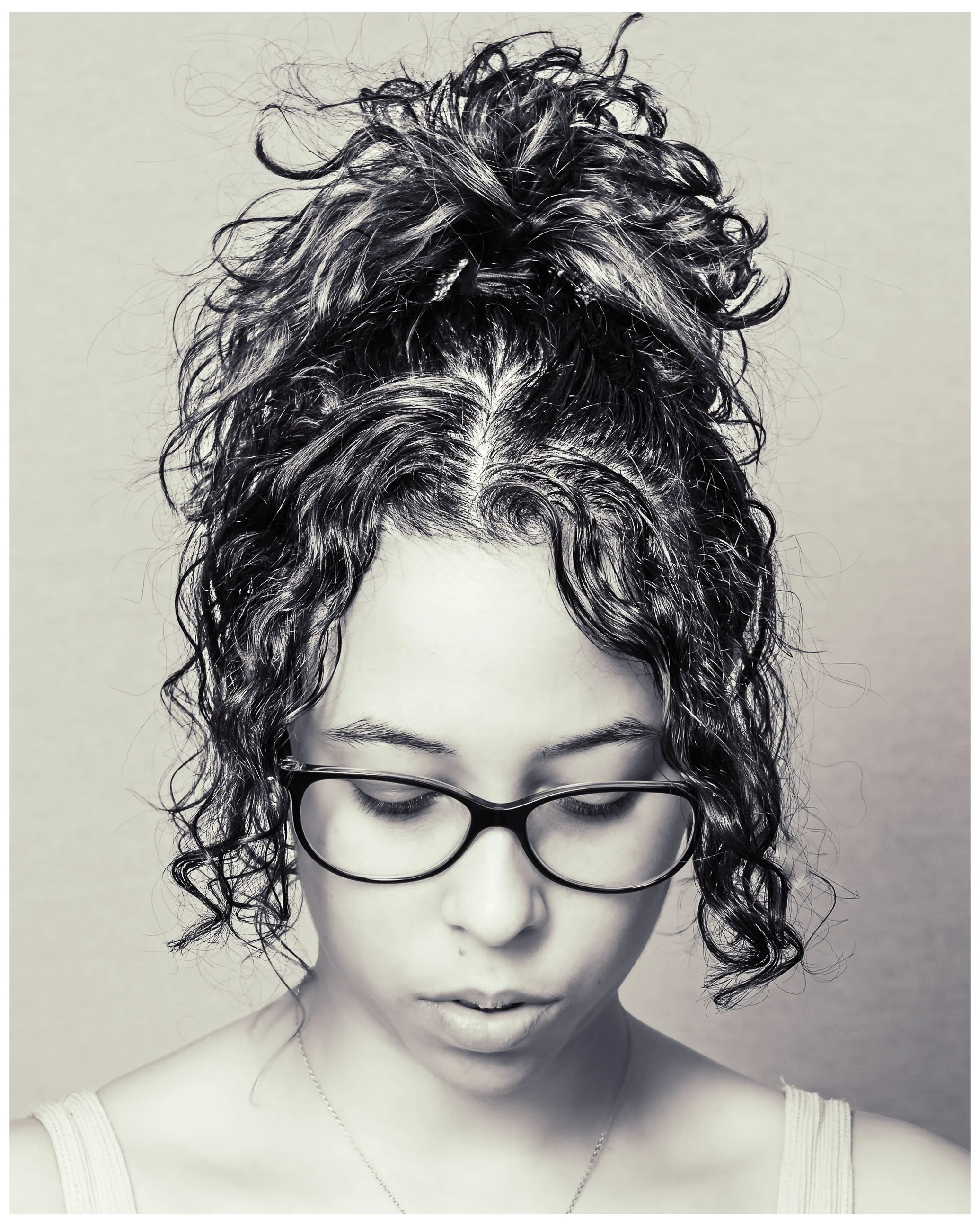 Black and white photo of a woman with curly hair styled in a high bun, wearing glasses, and looking downward.