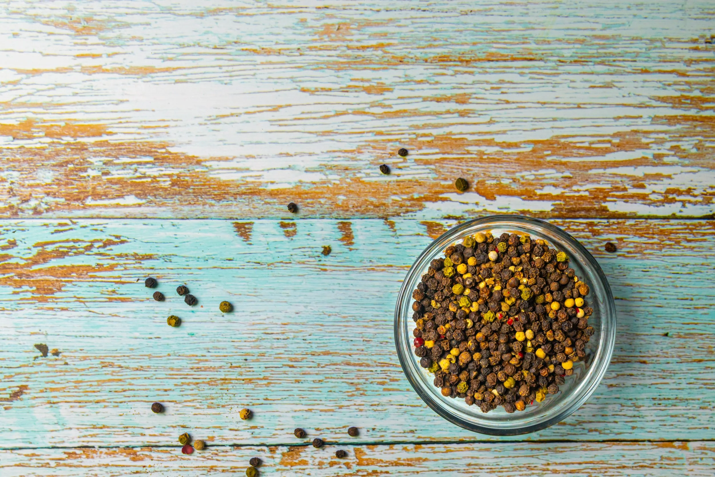 A glass bowl filled with mixed peppercorns on a rustic, painted wooden surface with scattered peppercorns around.