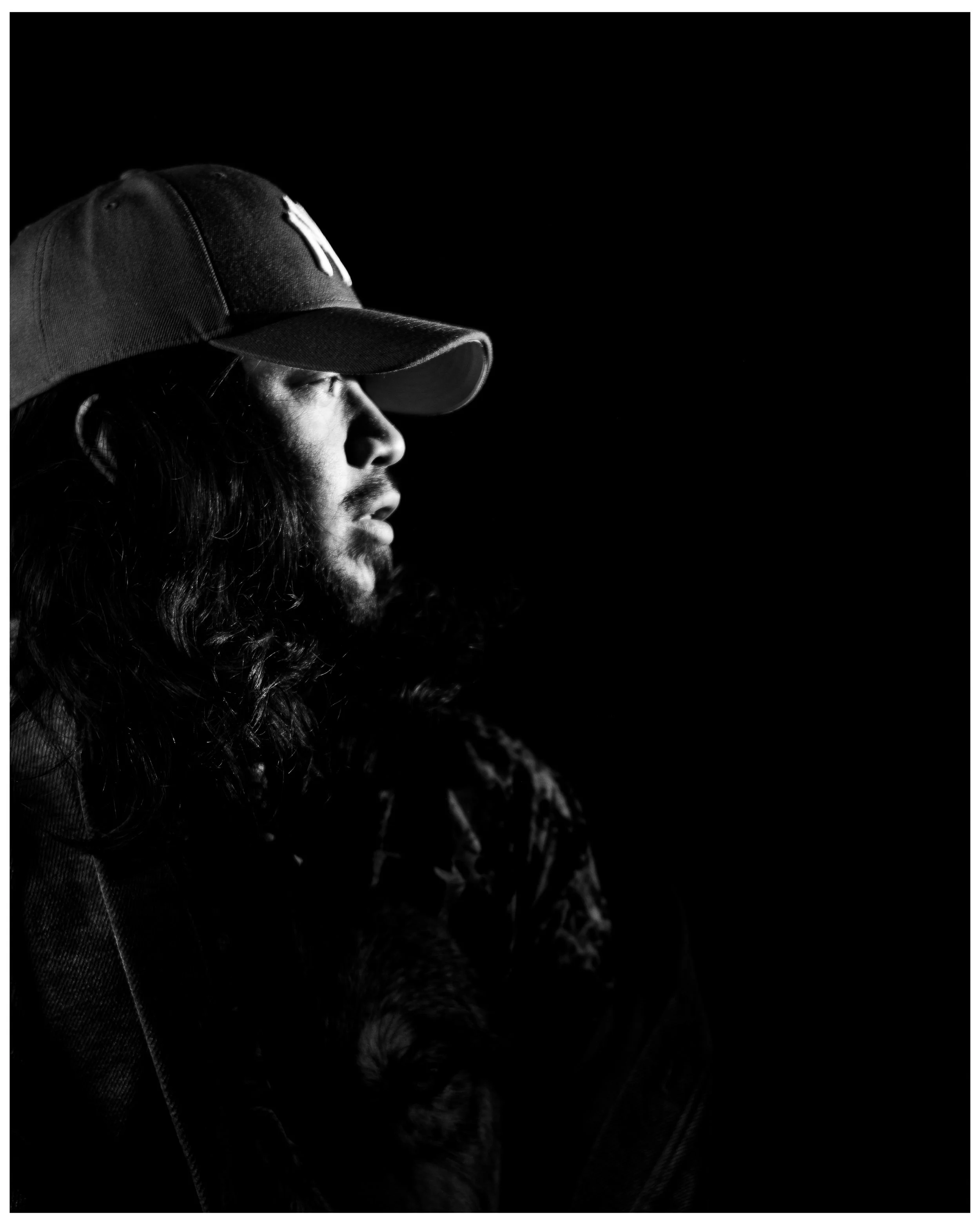 Black and white portrait of a man with long hair and beard wearing a baseball cap, looking to the side with a dark background.