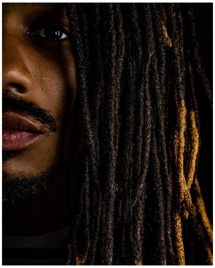 Close-up of half a man's face with dark skin, black textured beard, and long dreadlocks, with one side illuminated.