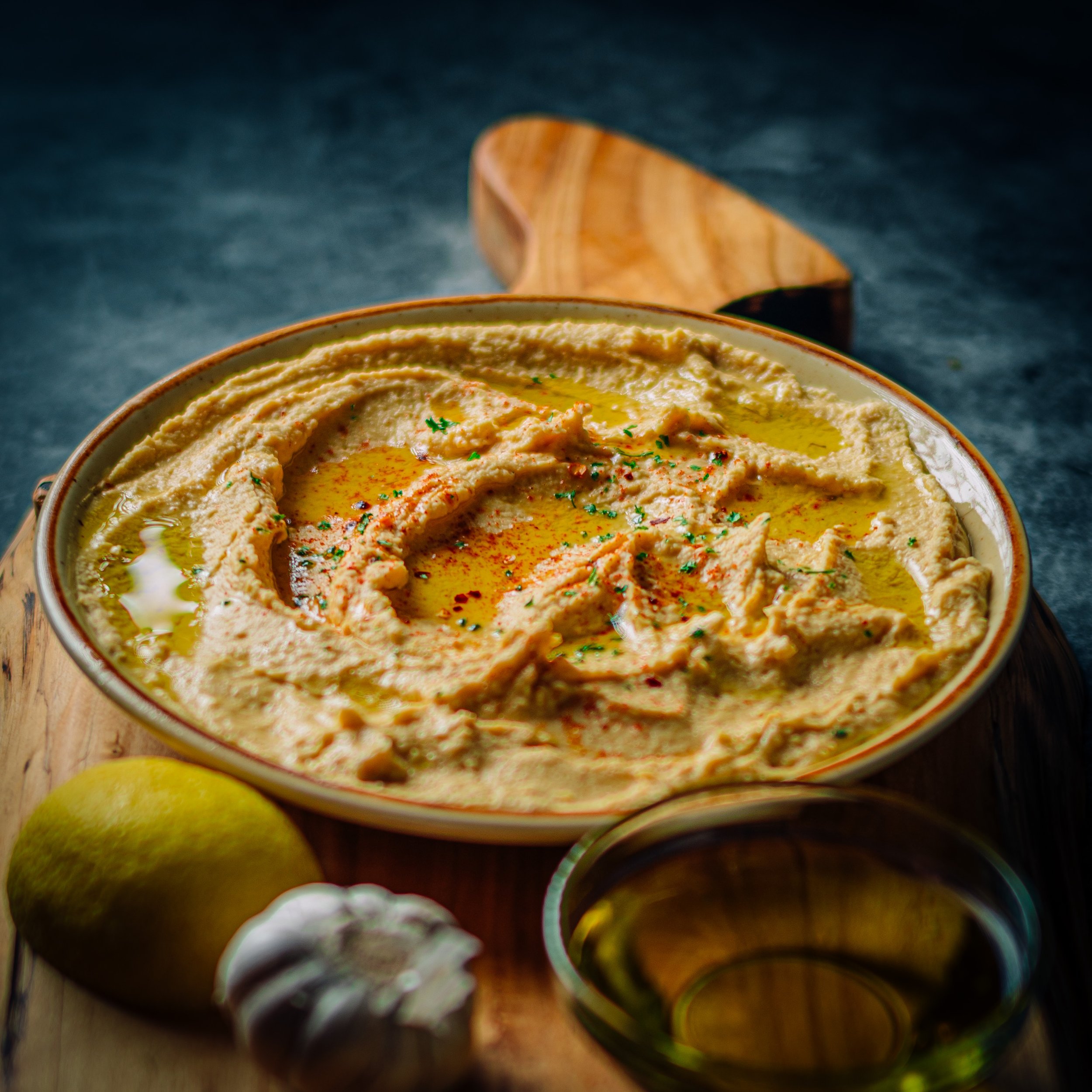 A bowl of hummus topped with olive oil and spices, placed on a wooden board with a lemon and garlic nearby, and a small bowl of olive oil in front.