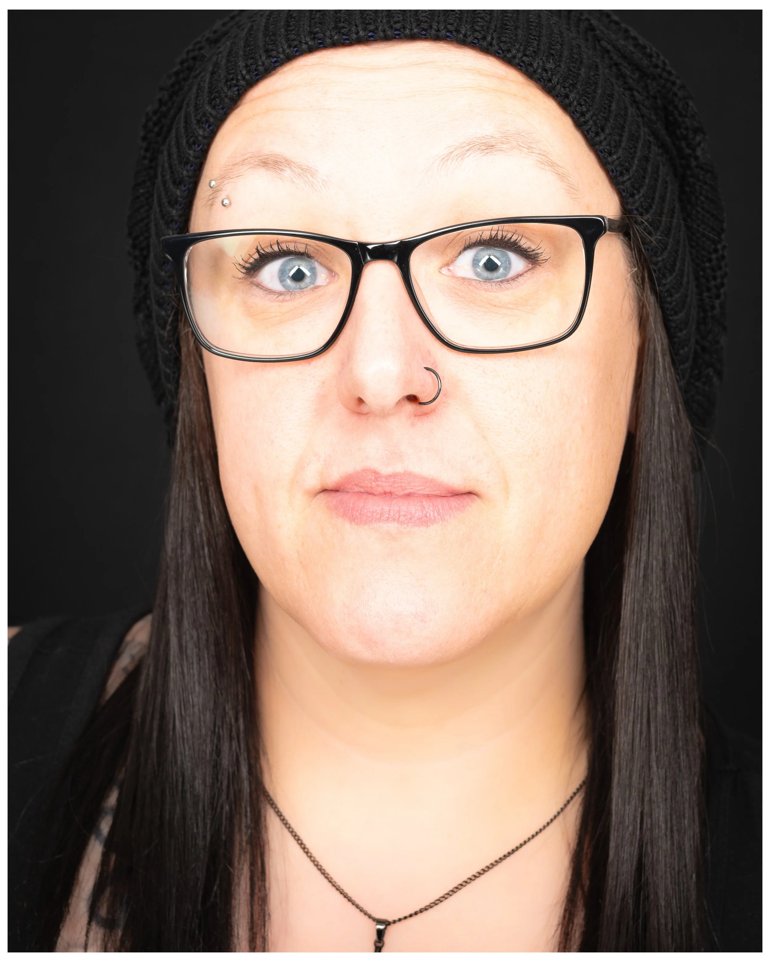 Close-up of a woman with light skin, blue eyes, wearing black glasses, a black knit beanie, and a small nose ring. She has long dark hair, a piercing above her right eyebrow, and a necklace with a small pendant.