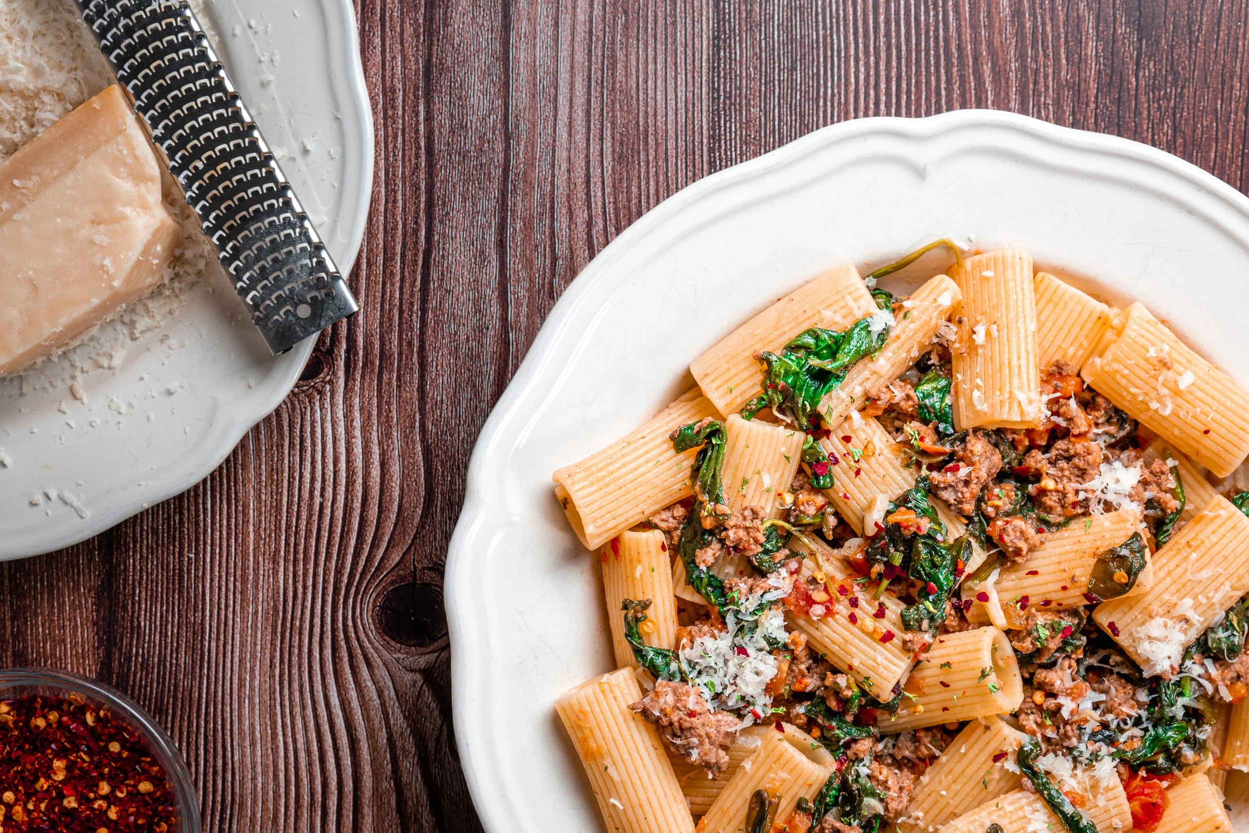 Pasta with ground meat, spinach, and grated cheese in a white dish on a wooden table, with a block of Parmesan cheese and a cheese grater nearby.