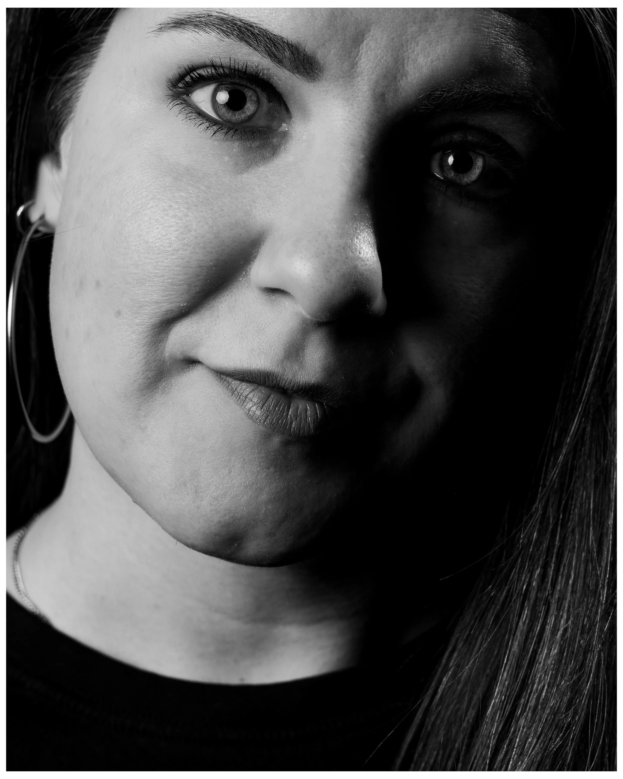 Close-up black and white photograph of a woman's face, with one eye illuminated and the other shadowed, showing detailed skin texture, eye makeup, and long hair.