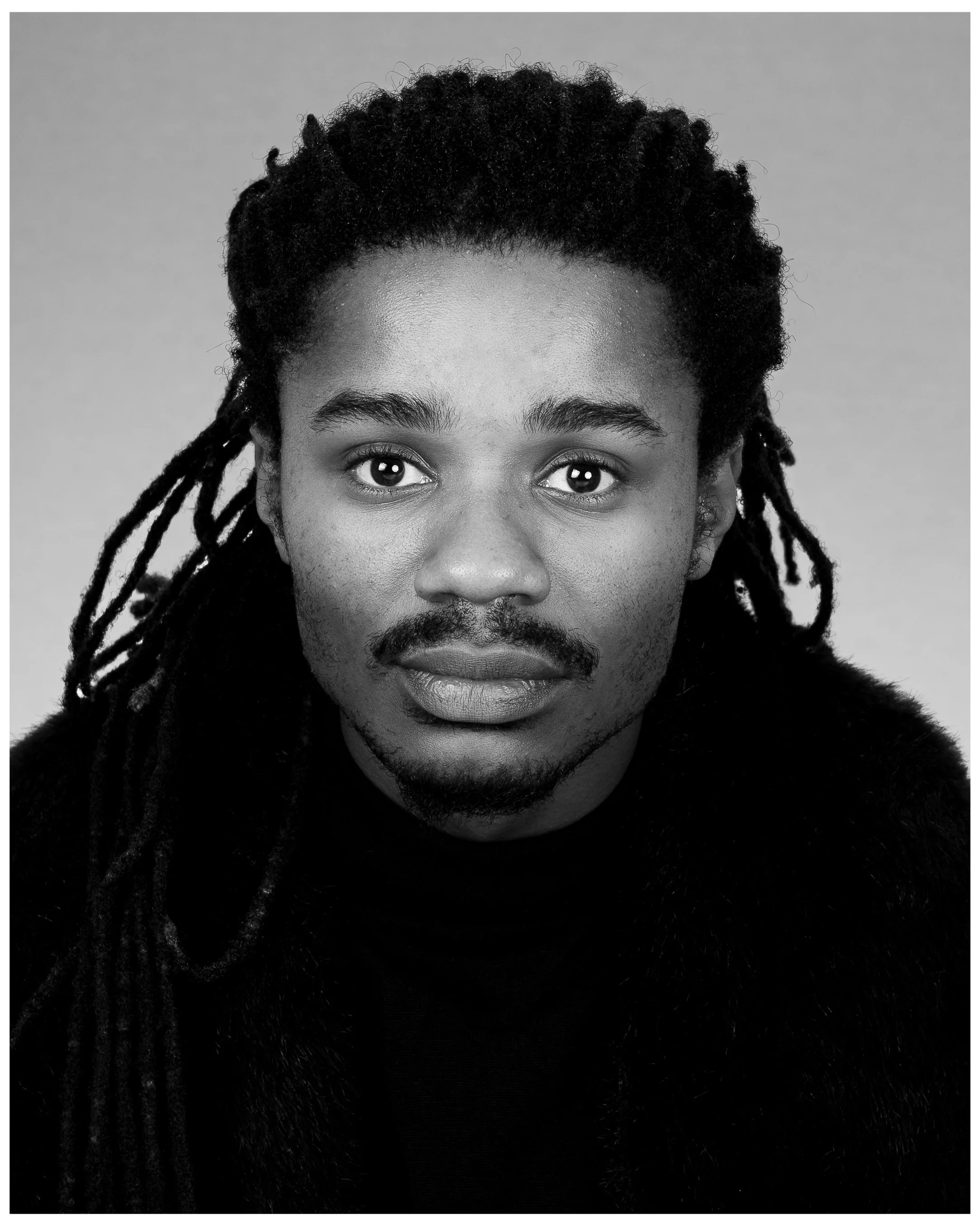 Black and white portrait of a young man with dreadlocks, a faint mustache, and a goatee, wearing a dark fur-collared garment, looking directly at the camera.