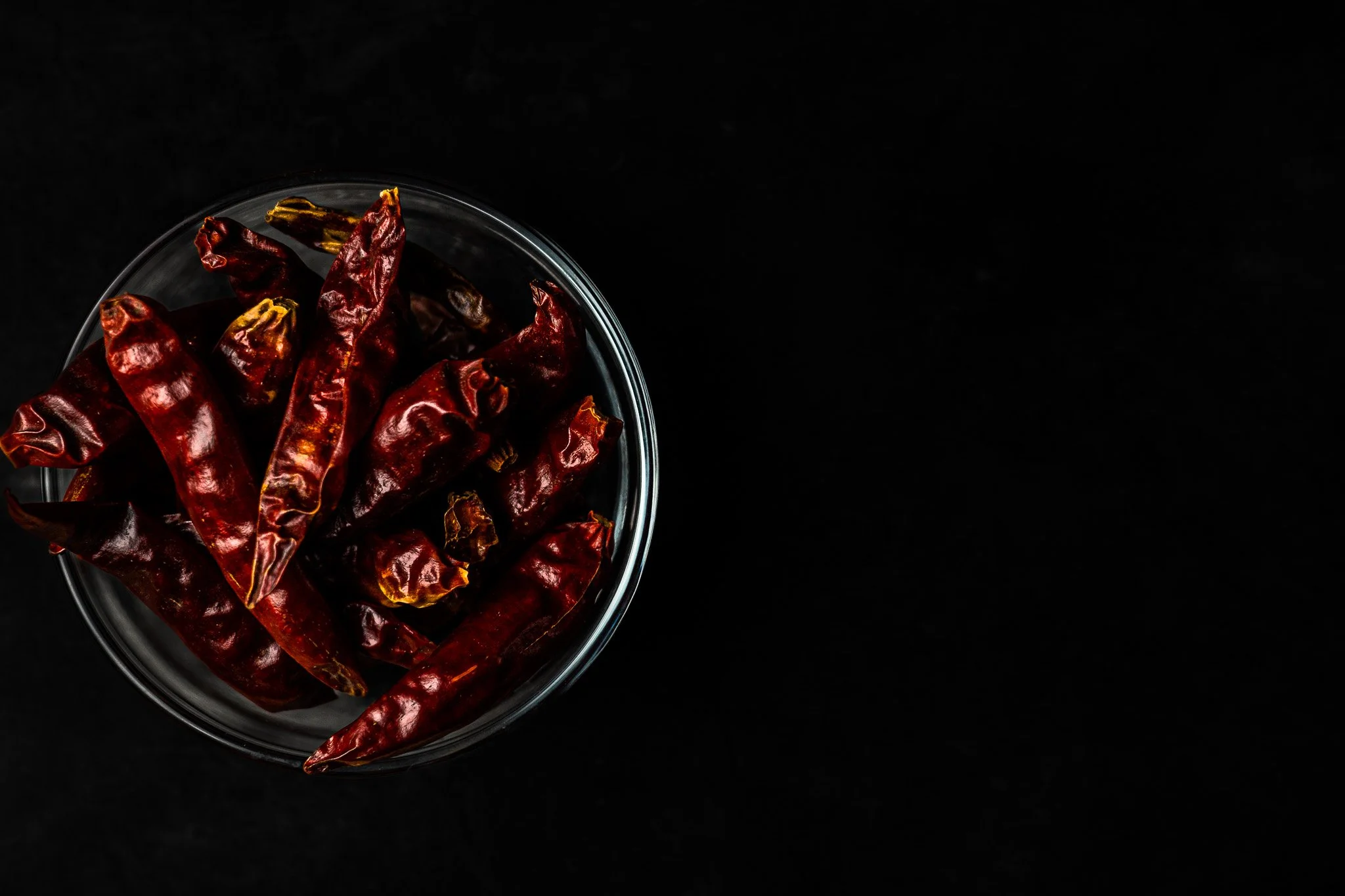 Red dried chili peppers in a glass bowl against a black background.