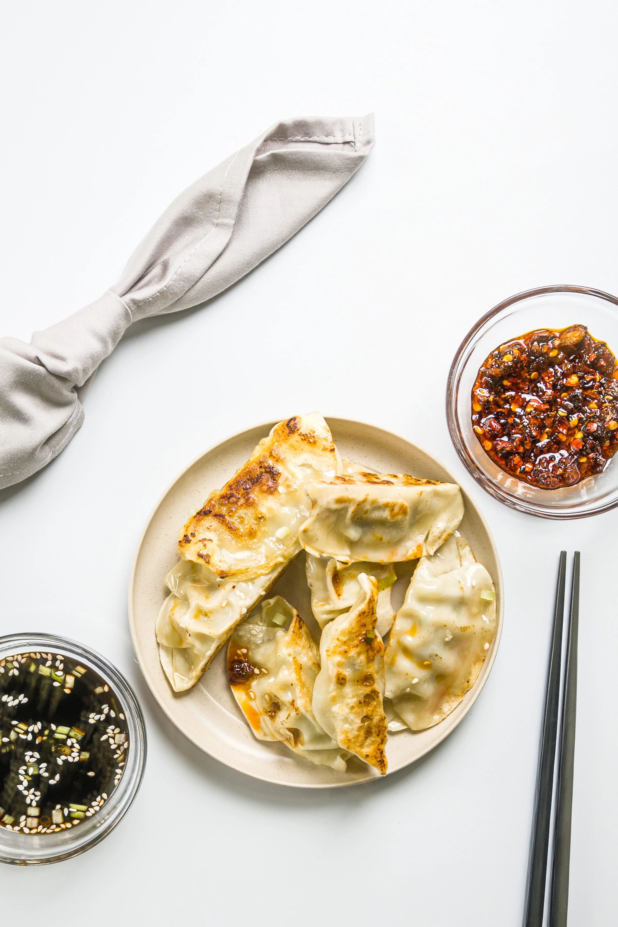 A plate of cooked dumplings with dipping sauces, a bowl of soy sauce, and a bowl of chili sauce, with chopsticks on a white background.