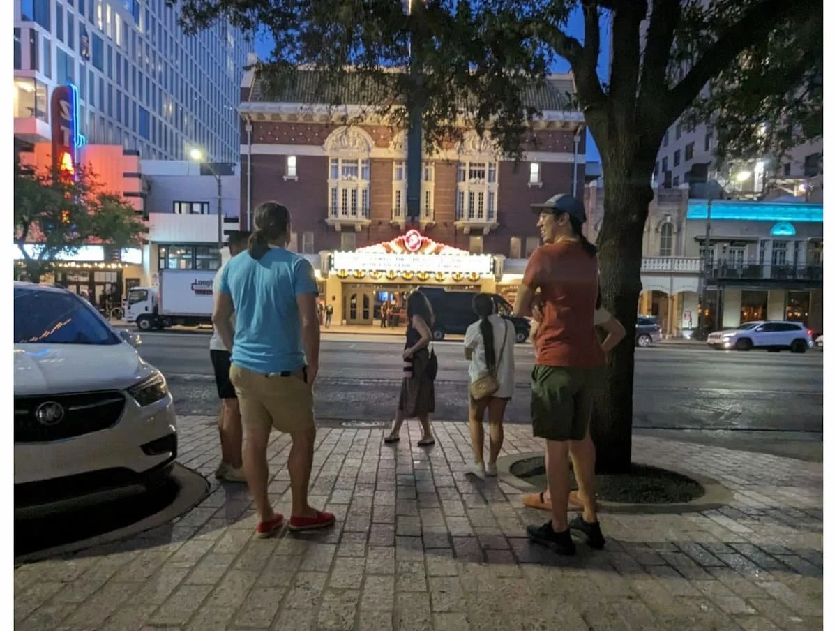 People standing on Congress Avenue sidewalk at dusk, with a lit Paramount theater in the background and cars driving by.
