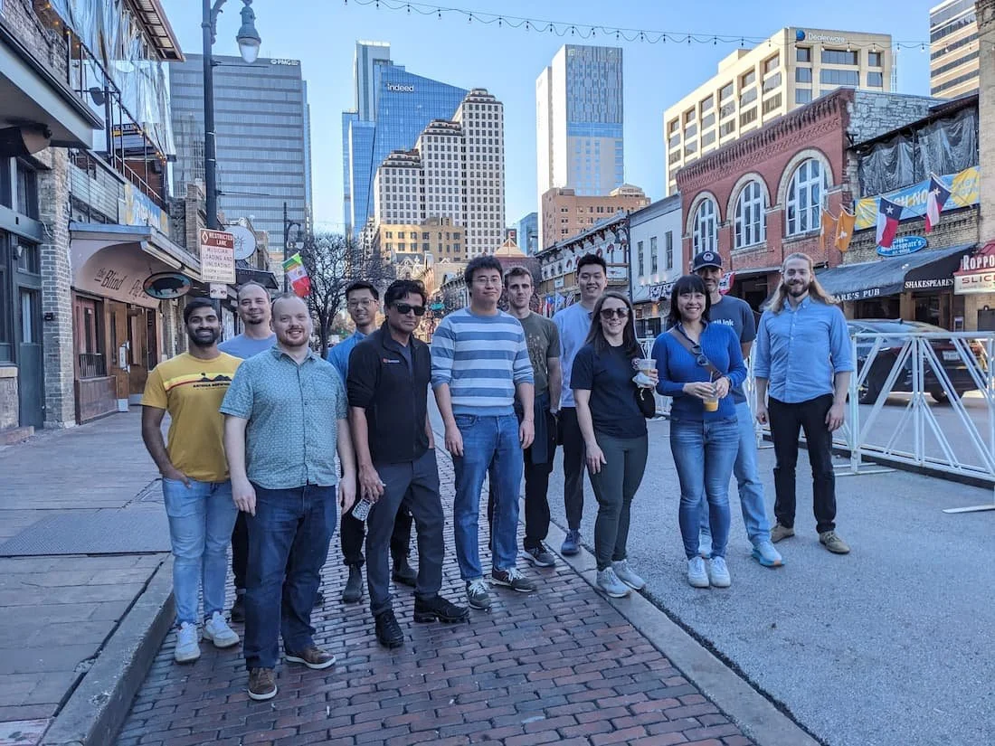 Group of people standing on a 6th street with tall buildings in the background, some smiling and holding drinks, during daytime.