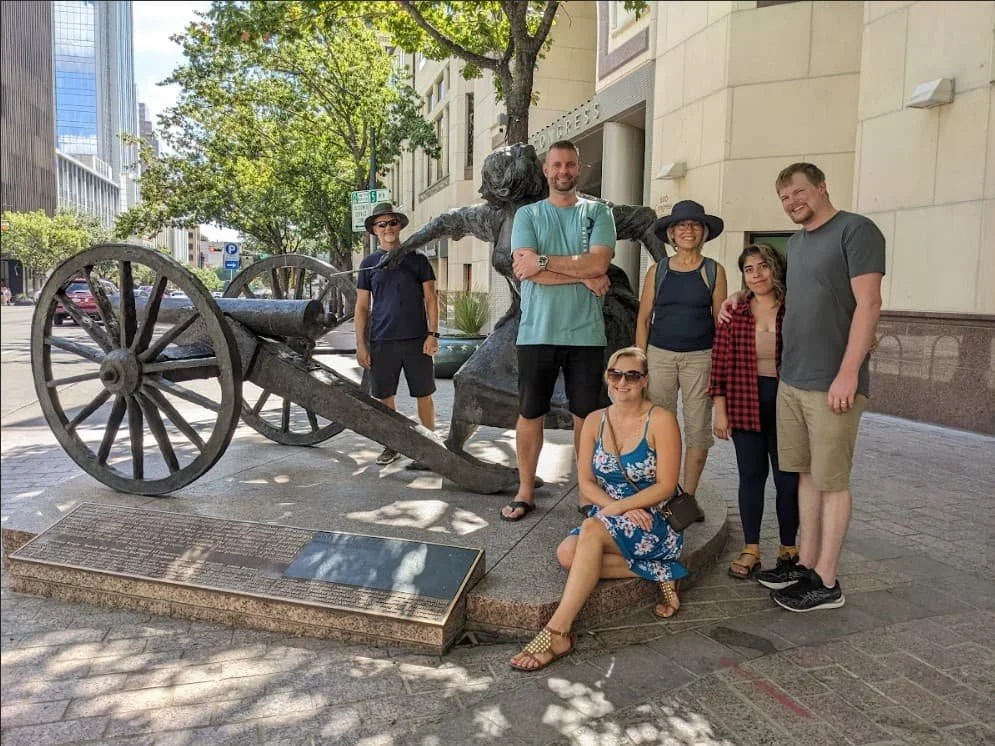Group of six people posing around a bronze statue of Angelina Eberly with a cannon, on Congress Avenue in Austin with trees and tall buildings in the background.