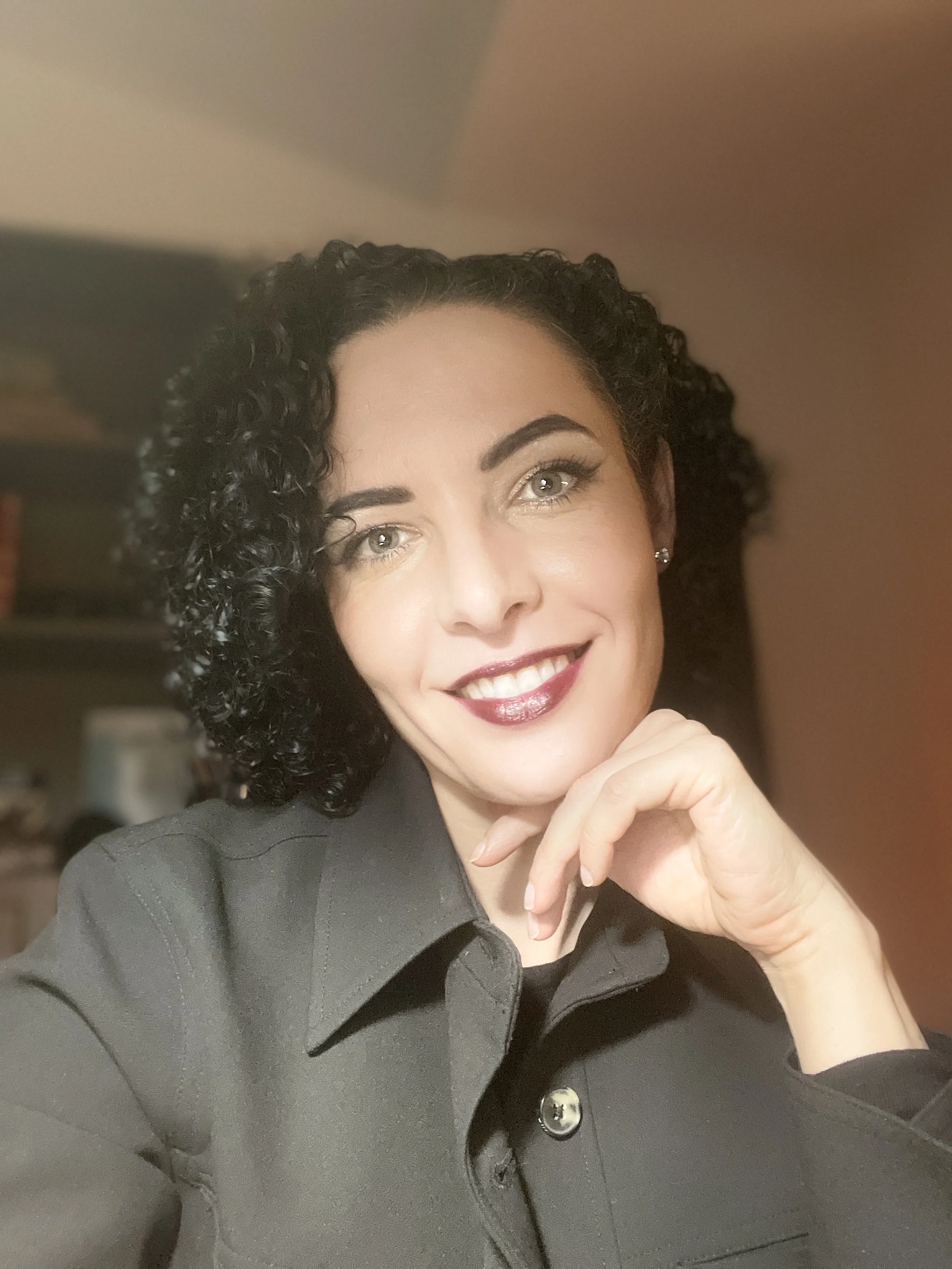 A woman with black curly hair and fair skin smiling at the camera, wearing a dark-colored shirt, in an indoor setting.