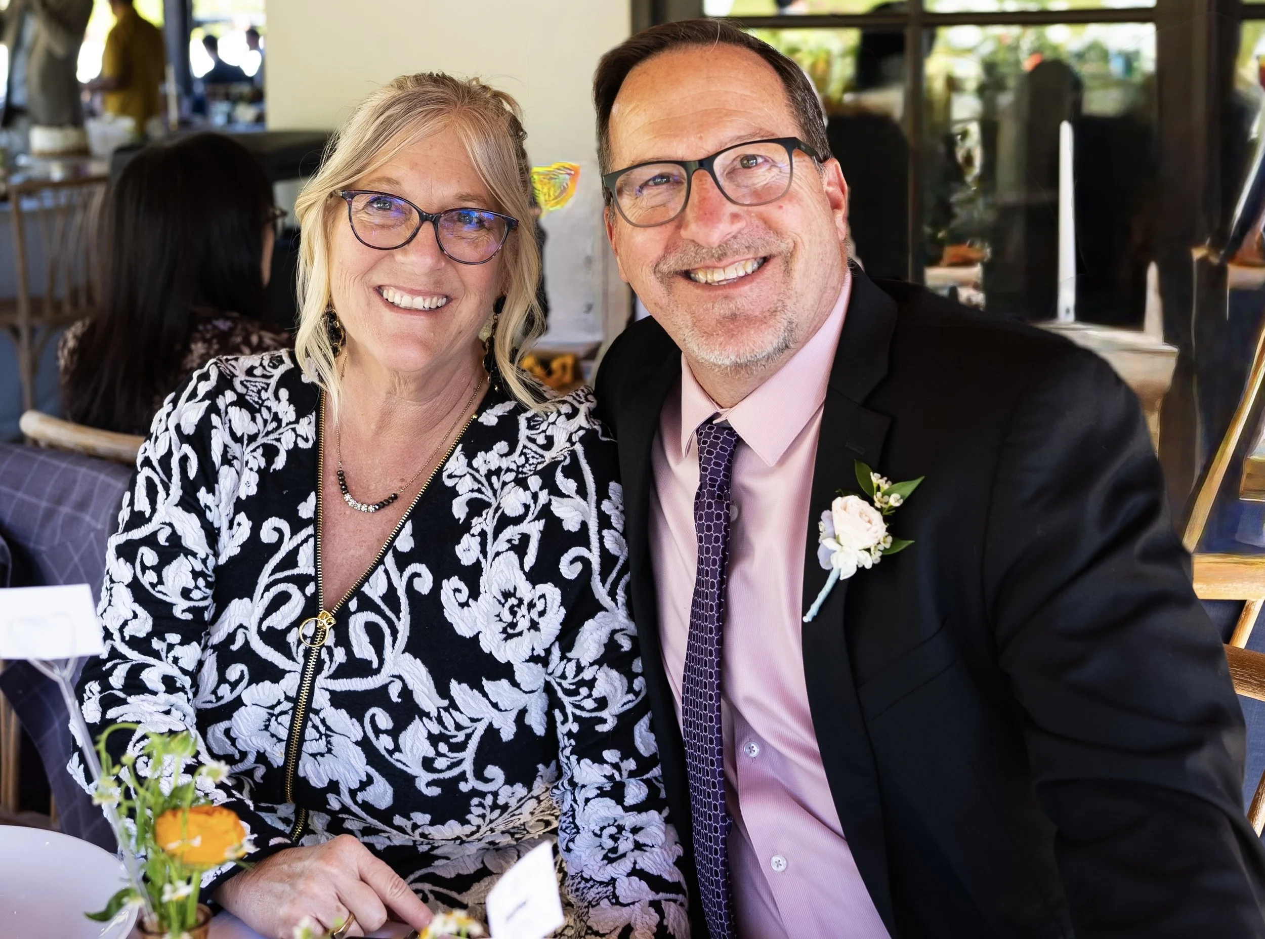 A smiling elderly woman with glasses and blonde hair, and a smiling man with glasses and dark hair, dressed in formal attire, sitting together at a celebration or wedding event with floral decorations and other guests in the background.