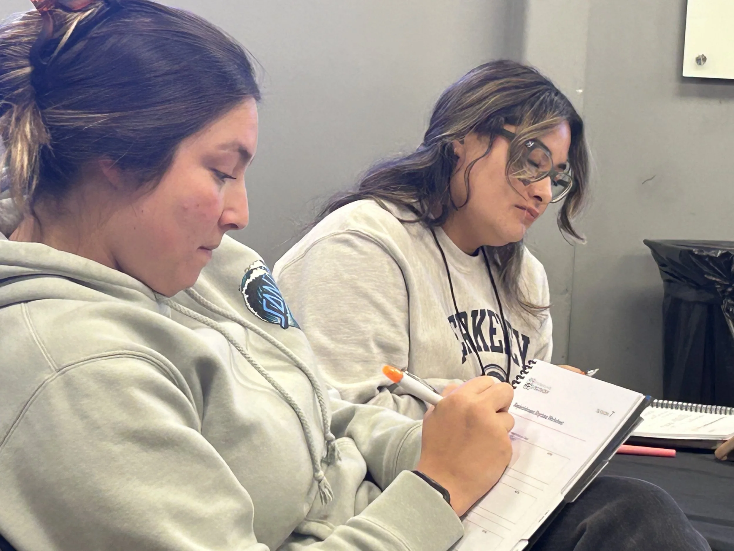 Two women sitting next to each other, reading notebooks or textbooks, with a gray wall in the background.