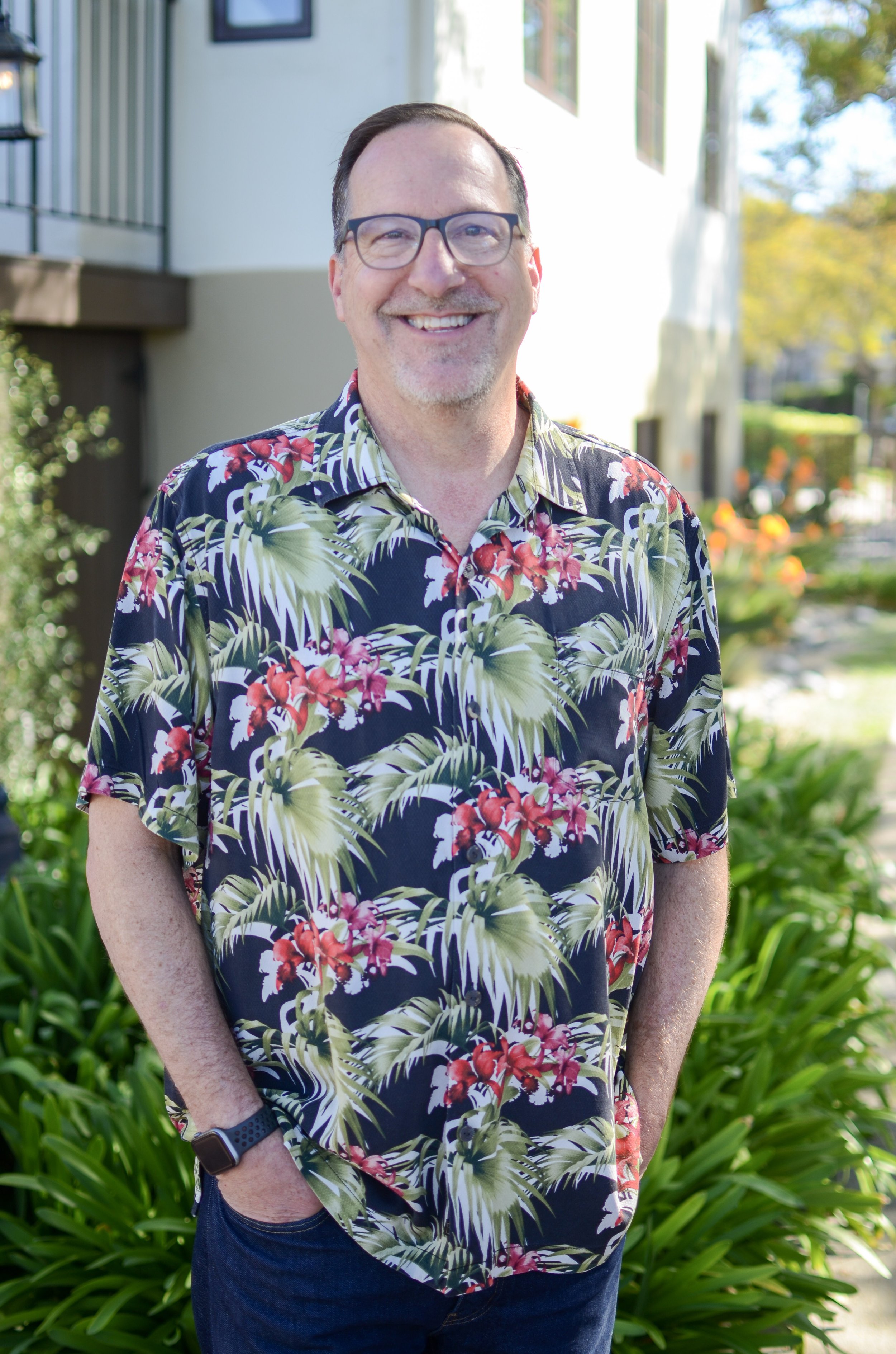 A middle-aged man smiling outdoors, wearing glasses, a colorful Hawaiian shirt with floral patterns, and a smartwatch on his left wrist. There are greenery and houses in the background on a sunny day.