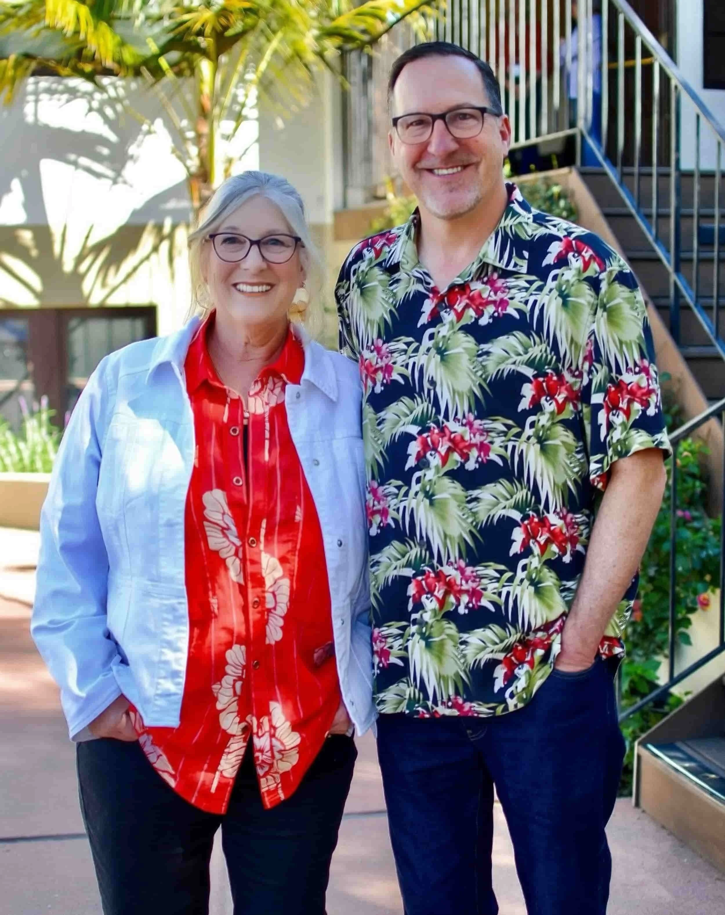 A smiling woman and man standing outdoors in front of a railing and plants, wearing casual clothes and glasses.
