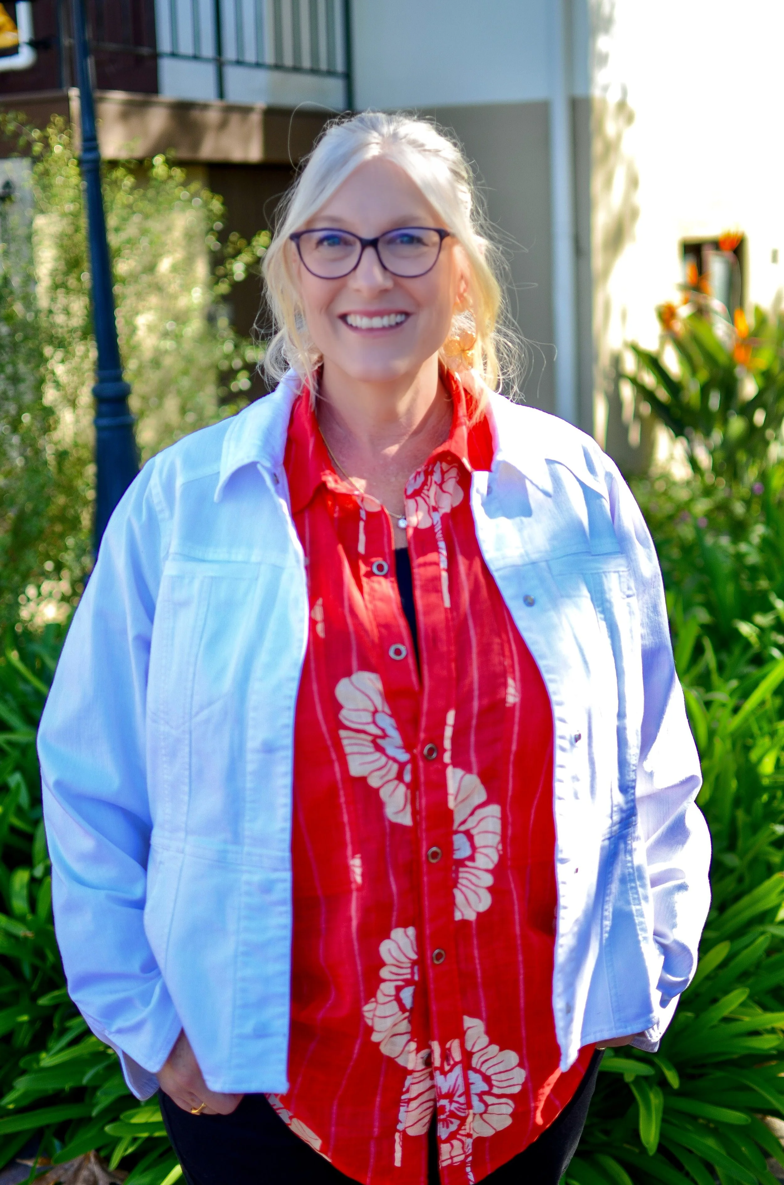 A smiling woman with glasses, wearing a red floral shirt and a white jacket, standing outdoors with greenery and a building in the background.