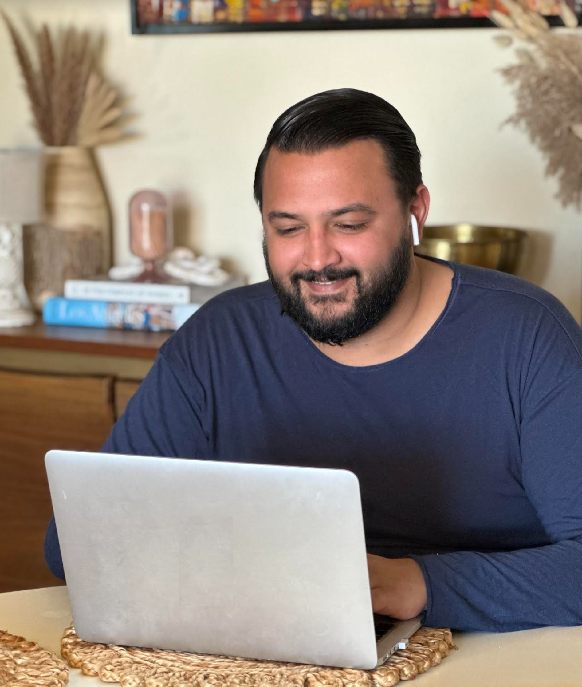A man with dark hair and a beard is sitting at a table, engaging in telehealth therapy on a laptop. He is wearing white wireless earbuds and a navy blue shirt. The background features books, decorative items, and a framed picture on the wall.