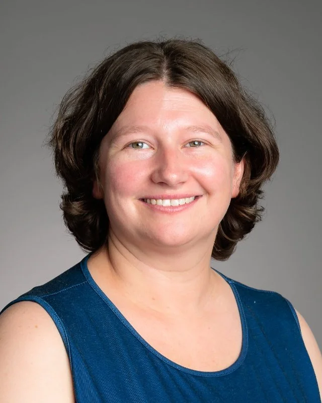 A woman with brown hair, glasses, and a gray T-shirt, smiling in front of a plain light gray background.