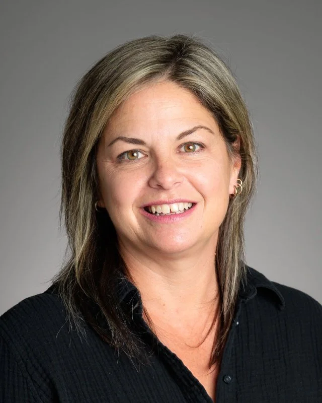 Headshot of a woman with shoulder-length light brown hair, wearing a black V-neck top and a gold necklace, smiling against a light gray background.
