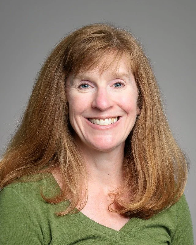 A woman with long red hair smiling, wearing a teal shirt, against a light gray background.
