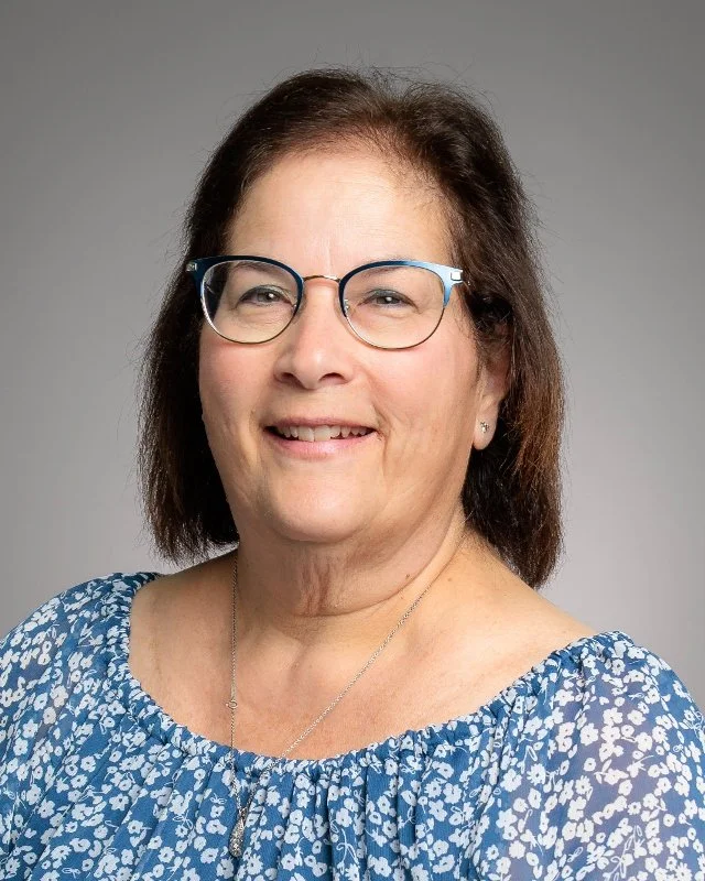 A woman with shoulder-length dark hair and glasses, smiling, wearing a sleeveless light blue top and two necklaces, against a plain gray background.
