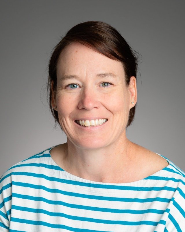Portrait of a woman with short light brown hair, wearing a red and navy striped shirt, smiling against a light gray background.