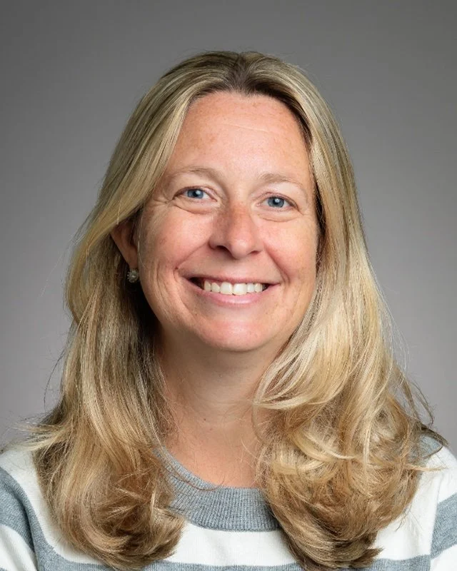 A woman with long blonde hair, blue eyes, and a bright smile wearing a red top against a light gray background.
