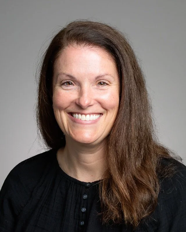 A woman with long brown hair, smiling, wearing a dark green top, against a plain gray background.