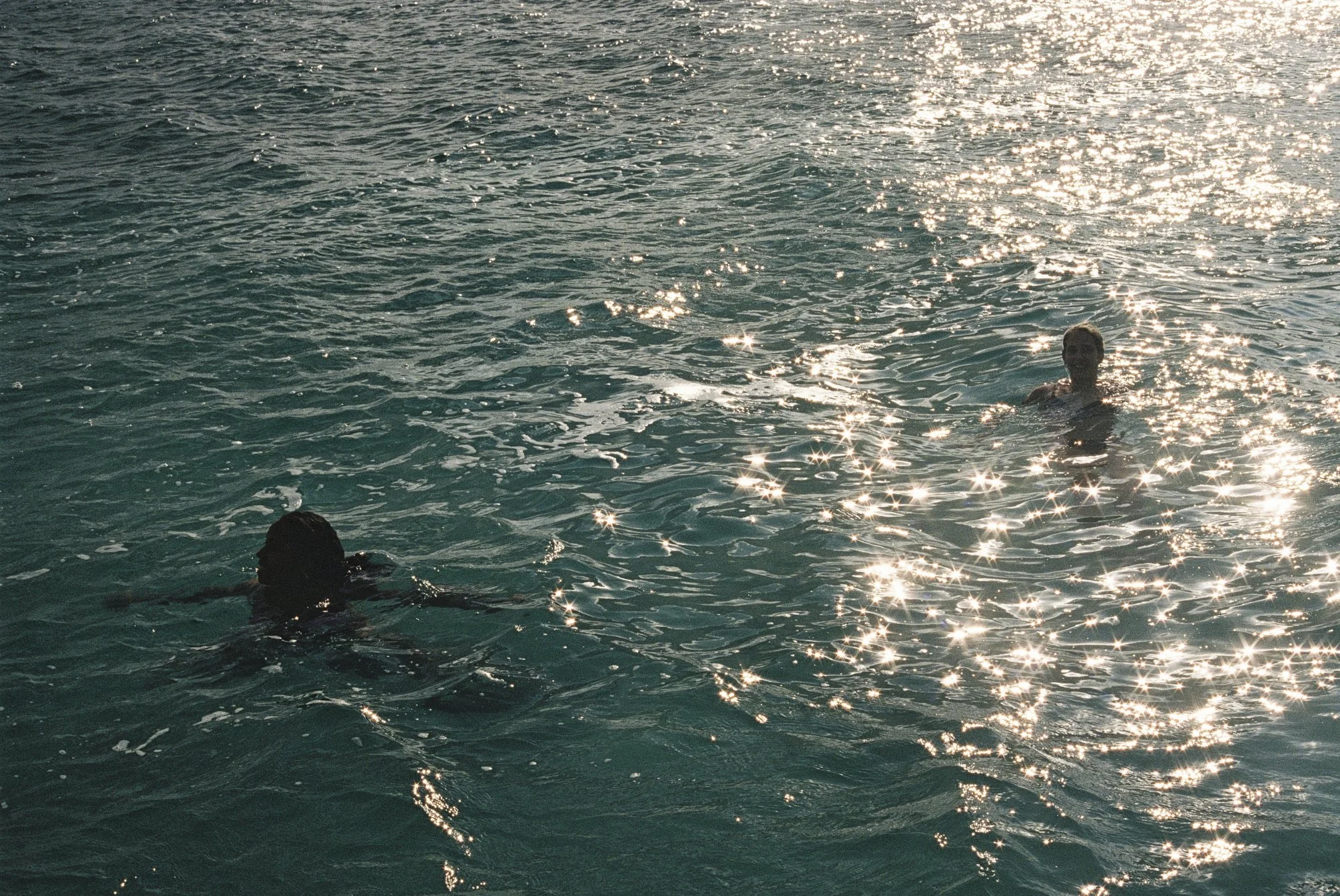 Two retreat participants are swimming in calm water, sunlight glinting on the surface, surrounded by nature.