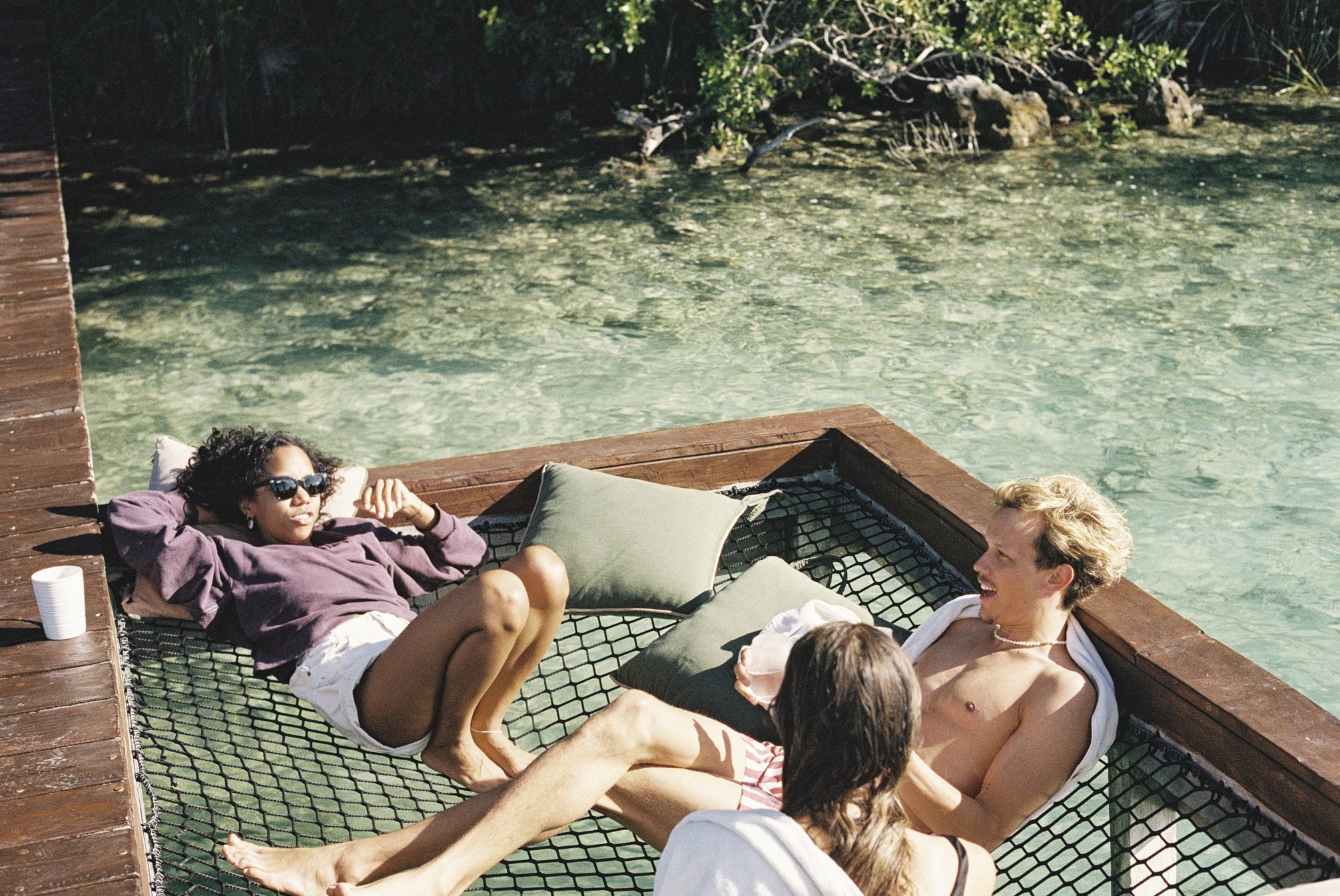 Three friends are relaxing on a floating netted platform in clear water at a retreat, surrounded by a wooden deck and trees in the background, capturing connection.