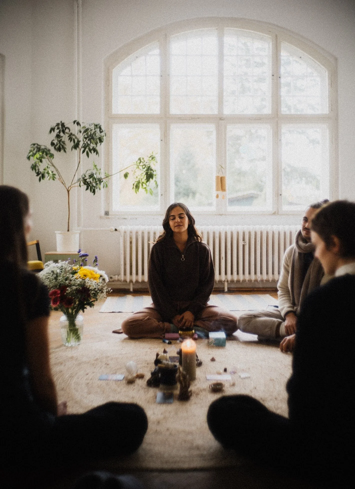 A group of people in a retreat sitting cross-legged on the floor of a bright, airy room with a large window, engaging in a meditation or coaching session. There are candles, crystals, and cards in front of them, with a vase of flowers on the side.