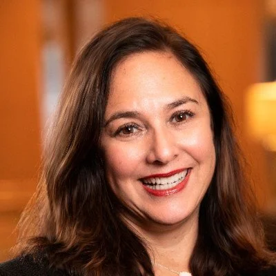 A smiling woman with long brown hair, wearing a black top, in an indoor setting with warm lighting.