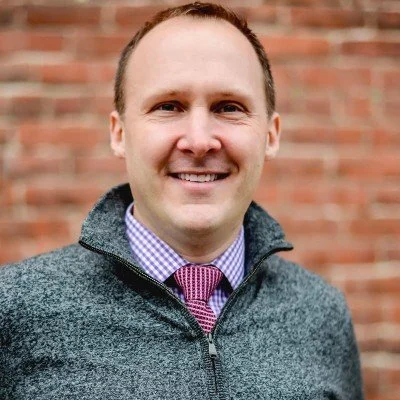 A smiling man wearing a gray quarter-zip sweater over a purple checkered shirt and pink tie, standing outdoors in front of a brick wall.