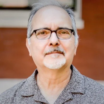 Portrait of a middle-aged man with gray hair, glasses, and a goatee, wearing a patterned shirt, standing outdoors with a brick building in the background.