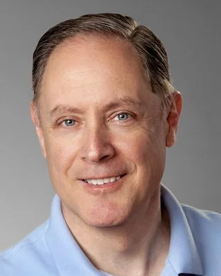 A headshot of a smiling man with light skin, short brown hair, and blue eyes, wearing a light blue collared shirt.